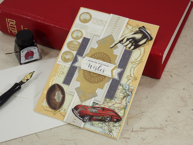 A handcrafted Birthday greeting card with gold foil details, a red car illustration, and a hand graphic, styled on a desk with a fountain pen, ink bottle, and red books.