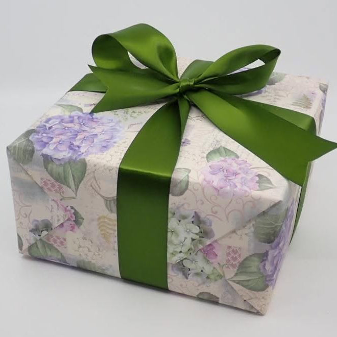 A square gift box wrapped in floral paper with purple flowers and green leaves, tied with a wide green satin ribbon and bow, on a light gray background.