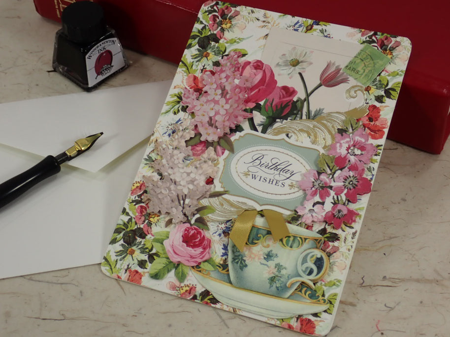 A handcrafted birthday card with a vintage floral design, featuring pink roses, a teal teacup, and a central label reading 'Birthday Wishes'. The card is styled on a marble surface next to a fountain pen, a white envelope, and a red box.