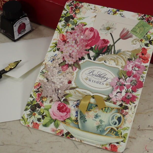 A handcrafted birthday card with a vintage floral design, featuring pink roses, a teal teacup, and a central label reading 'Birthday Wishes'. The card is styled on a marble surface next to a fountain pen, a white envelope, and a red box.