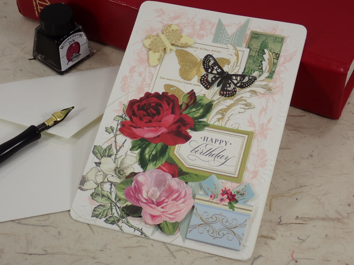 A handcrafted birthday greeting card with vintage-style floral and butterfly illustrations, placed on a marble-like surface next to a fountain pen, ink bottle, and red box.