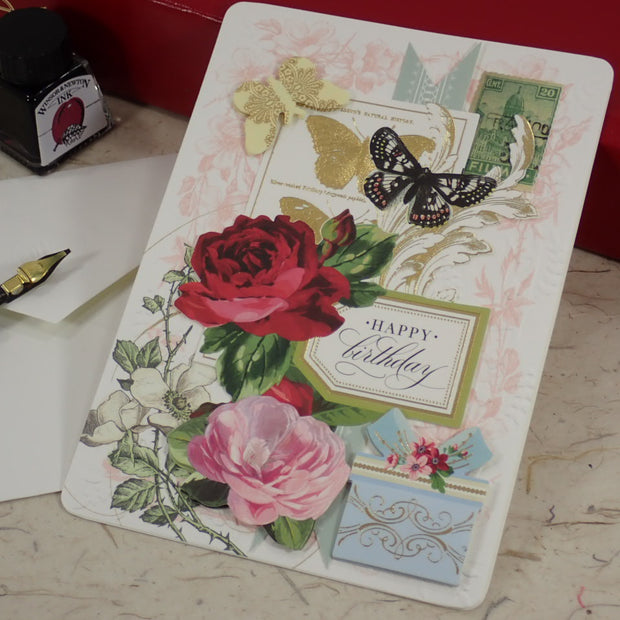 A handcrafted birthday greeting card with vintage-style floral and butterfly illustrations, placed on a marble-like surface next to a fountain pen, ink bottle, and red box.