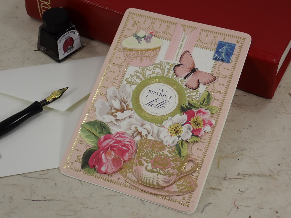 A handmade birthday greeting card with vintage floral and butterfly designs, placed on a tabletop next to a fountain pen, ink bottle, and red book.
