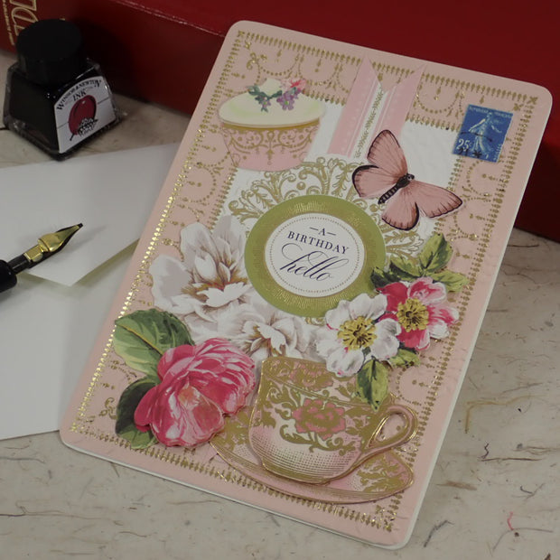 A handmade birthday greeting card with vintage floral and butterfly designs, placed on a tabletop next to a fountain pen, ink bottle, and red book.