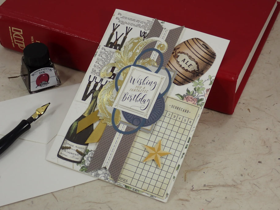 A handmade birthday card with vintage-inspired collage elements including a bottle label, barrel graphic, grid paper, and a blue circular frame reading 'Wishing you a Birthday.' The card is styled on a tabletop with a red book, ink bottle, and fountain pen nearby.
