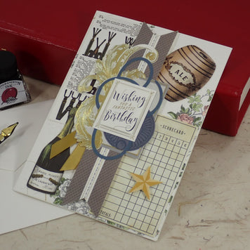 A handmade birthday card with vintage-inspired collage elements including a bottle label, barrel graphic, grid paper, and a blue circular frame reading 'Wishing you a Birthday.' The card is styled on a tabletop with a red book, ink bottle, and fountain pen nearby.