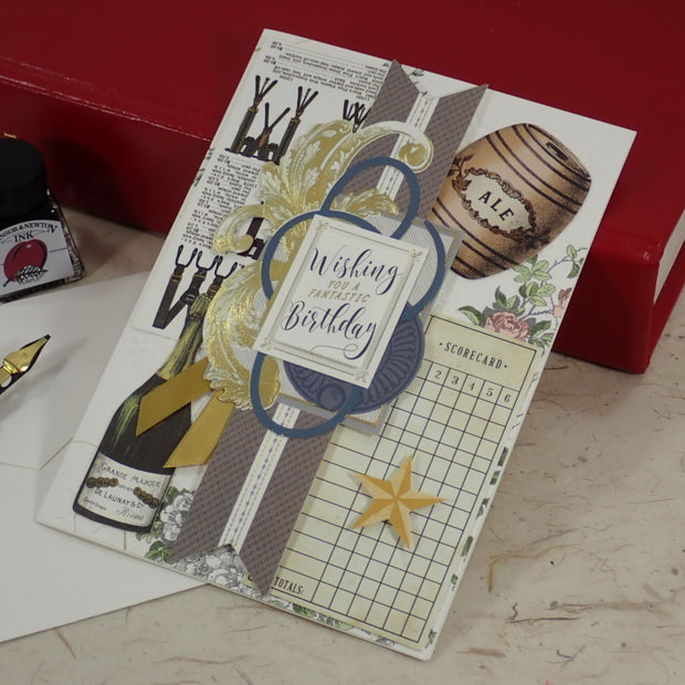 A handmade birthday card with vintage-inspired collage elements including a bottle label, barrel graphic, grid paper, and a blue circular frame reading 'Wishing you a Birthday.' The card is styled on a tabletop with a red book, ink bottle, and fountain pen nearby.