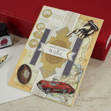 A handcrafted Birthday greeting card with gold foil details, a red car illustration, and a hand graphic, styled on a desk with a fountain pen, ink bottle, and red books.