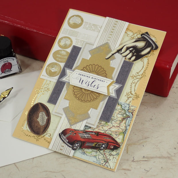 A handcrafted Birthday greeting card with gold foil details, a red car illustration, and a hand graphic, styled on a desk with a fountain pen, ink bottle, and red books.
