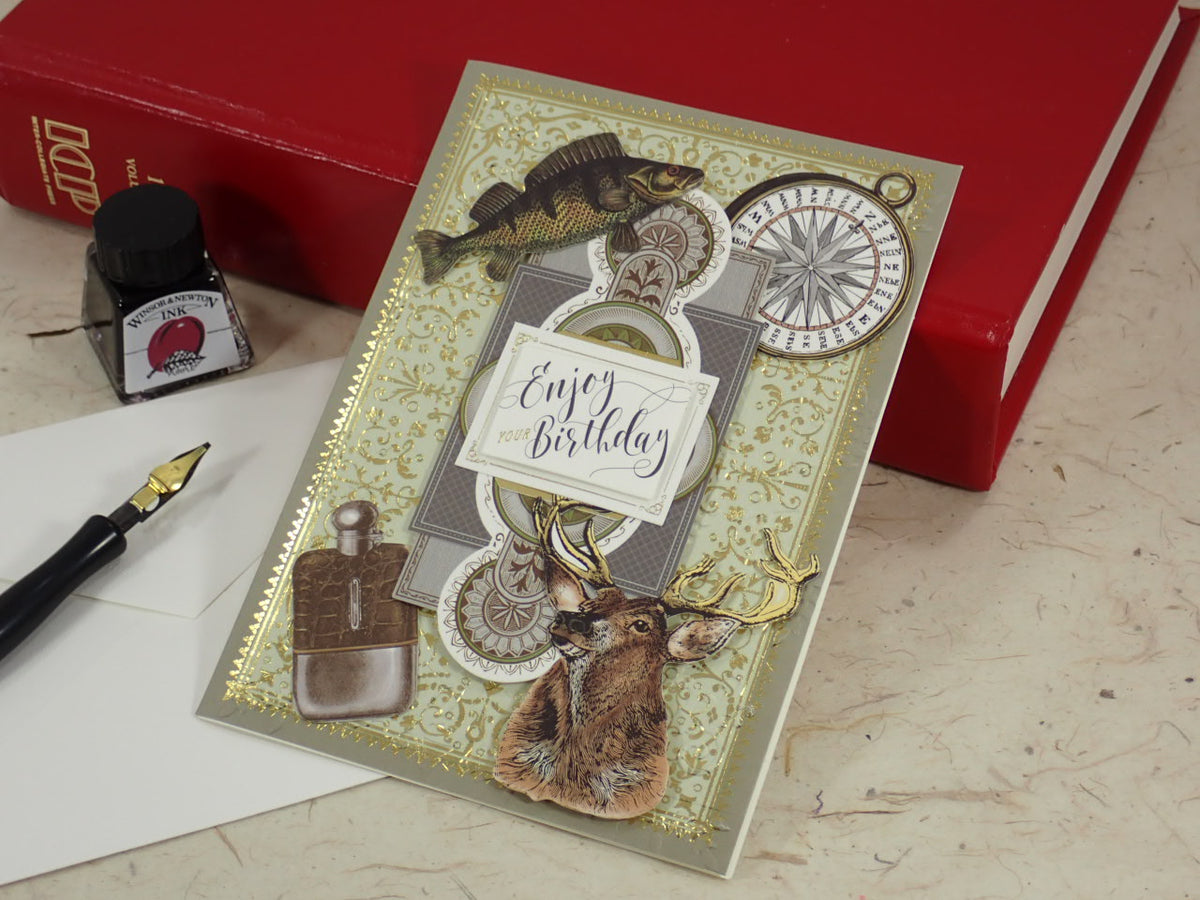 A handcrafted birthday card with vintage-style illustrations of a fish, compass, deer head, and a pocket watch, styled on a tabletop with a red book, fountain pen, and ink bottle.