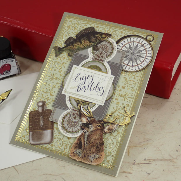 A handcrafted birthday card with vintage-style illustrations of a fish, compass, deer head, and a pocket watch, styled on a tabletop with a red book, fountain pen, and ink bottle.