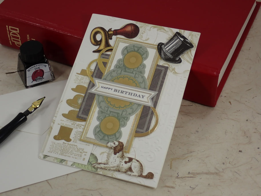 A handcrafted birthday greeting card with vintage-style illustrations, including a dog, top hat, gold ribbon, and 'HAPPY BIRTHDAY' banner, styled on a tabletop with a red book, ink bottle, and fountain pen.