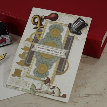 A handcrafted birthday greeting card with vintage-style illustrations, including a dog, top hat, gold ribbon, and 'HAPPY BIRTHDAY' banner, styled on a tabletop with a red book, ink bottle, and fountain pen.