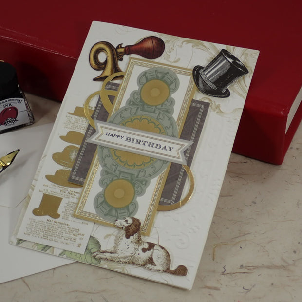 A handcrafted birthday greeting card with vintage-style illustrations, including a dog, top hat, gold ribbon, and 'HAPPY BIRTHDAY' banner, styled on a tabletop with a red book, ink bottle, and fountain pen.