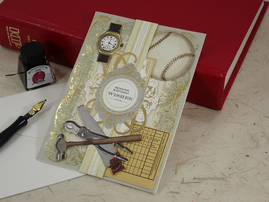 A baseball-themed handcrafted Birthday greeting card with gold foil details, a baseball graphic, a watch, and sports tools, styled on a desk next to a red book, fountain pen, and ink bottle.