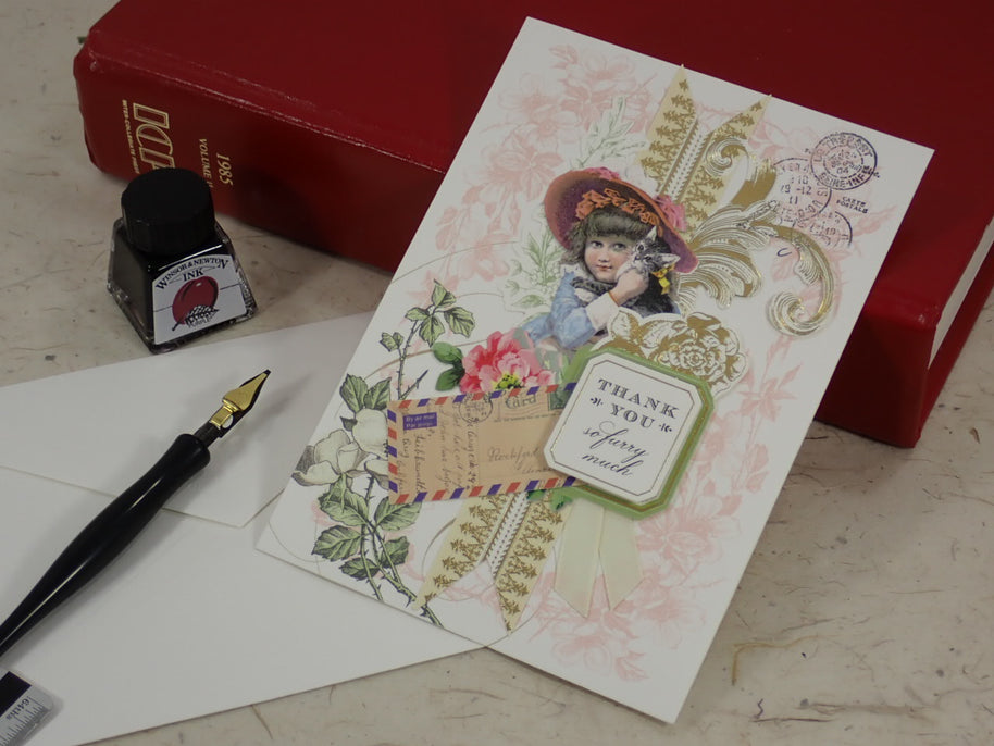 A handcrafted Thank You greeting card with a child in a hat, floral details, an envelope, a fountain pen, and a small ink bottle arranged on a light surface, with red books in the background.