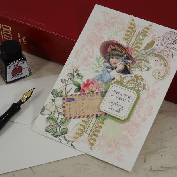 A handcrafted Thank You greeting card with a child in a hat, floral details, an envelope, a fountain pen, and a small ink bottle arranged on a light surface, with red books in the background.