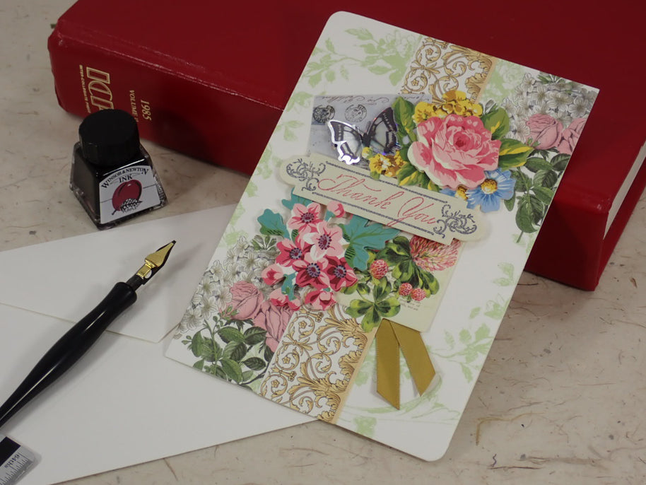 A handcrafted Thank You greeting card with floral and ornate collage designs, shown standing upright on a tabletop with an envelope, fountain pen, and red books in the background. 