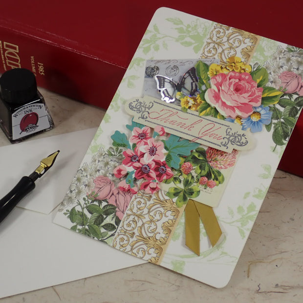 A handcrafted Thank You greeting card with floral and ornate collage designs, shown standing upright on a tabletop with an envelope, fountain pen, and red books in the background. 