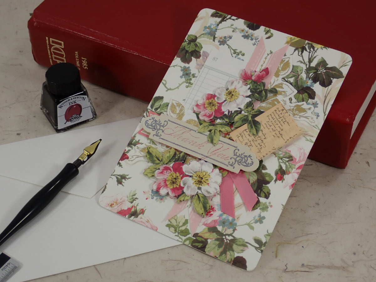 A handcrafted Thank You greeting card with pink ribbons and a small hanging tag, styled on a tabletop with a fountain pen, ink bottle, white envelope, and red books in the background.