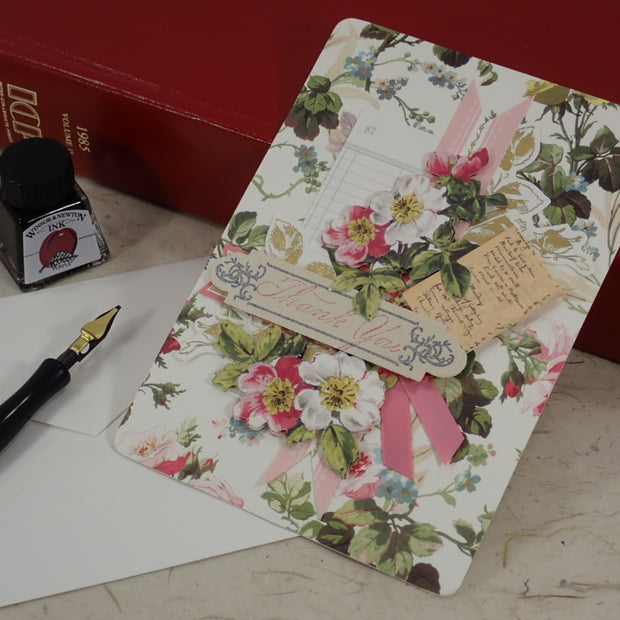 A handcrafted Thank You greeting card with pink ribbons and a small hanging tag, styled on a tabletop with a fountain pen, ink bottle, white envelope, and red books in the background.