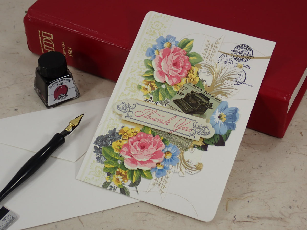 A handcrafted Thank You card with layered floral artwork, postage stamp graphics, and a central 'Thank You' banner, styled on a tabletop with a fountain pen, ink bottle, white envelopes, and red books.