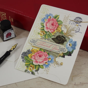 A handcrafted Thank You card with layered floral artwork, postage stamp graphics, and a central 'Thank You' banner, styled on a tabletop with a fountain pen, ink bottle, white envelopes, and red books.