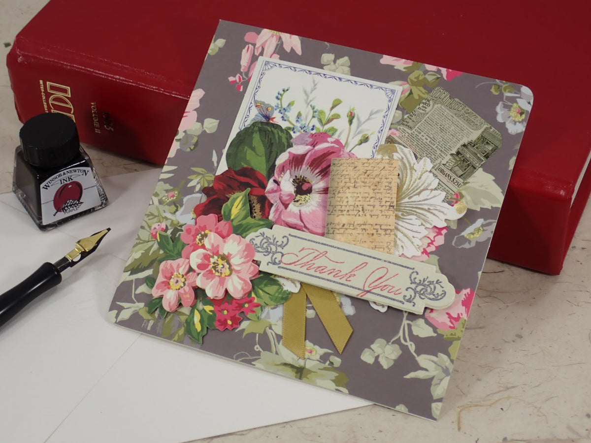 A handcrafted floral thank-you card with pink flowers, vintage script, and a gold ribbon bow, styled on a marble surface with a red box, ink bottle, and fountain pen nearby.
