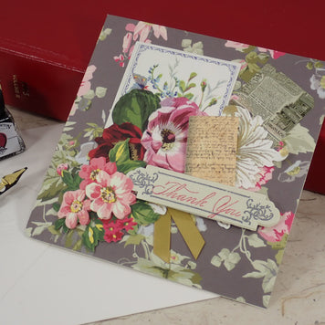 A handcrafted floral thank-you card with pink flowers, vintage script, and a gold ribbon bow, styled on a marble surface with a red box, ink bottle, and fountain pen nearby.