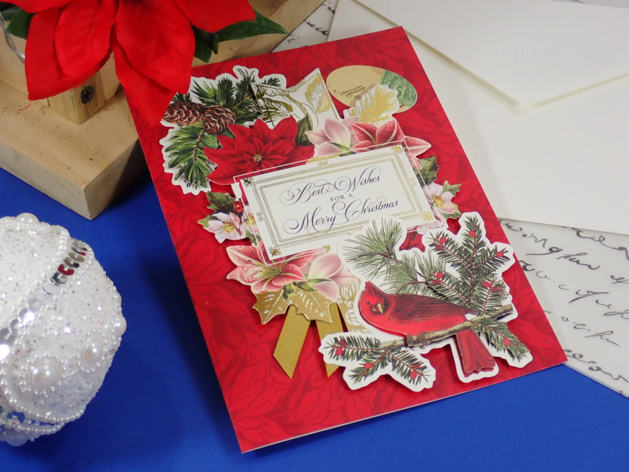 A handcrafted, red Christmas greeting card with layered cutout decorations including a red cardinal, poinsettias, pine branches, a gold bow, and a central label reading 'Best Wishes for a Merry Christmas.' 