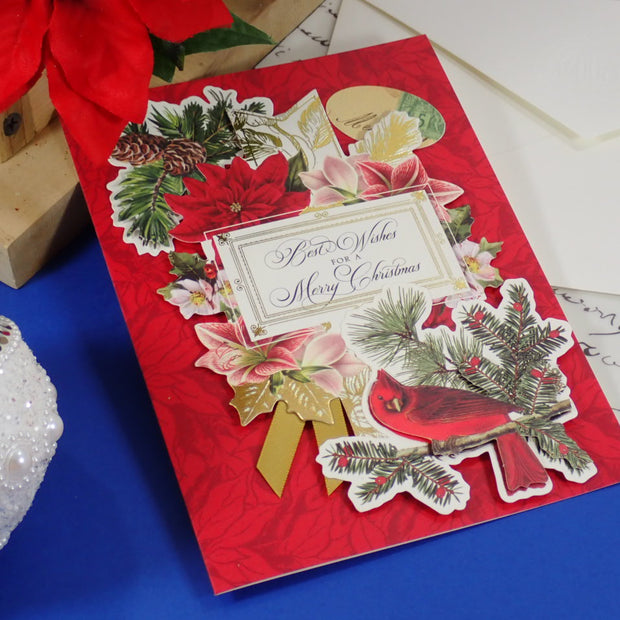 A handcrafted, red Christmas greeting card with layered cutout decorations including a red cardinal, poinsettias, pine branches, a gold bow, and a central label reading 'Best Wishes for a Merry Christmas.' 