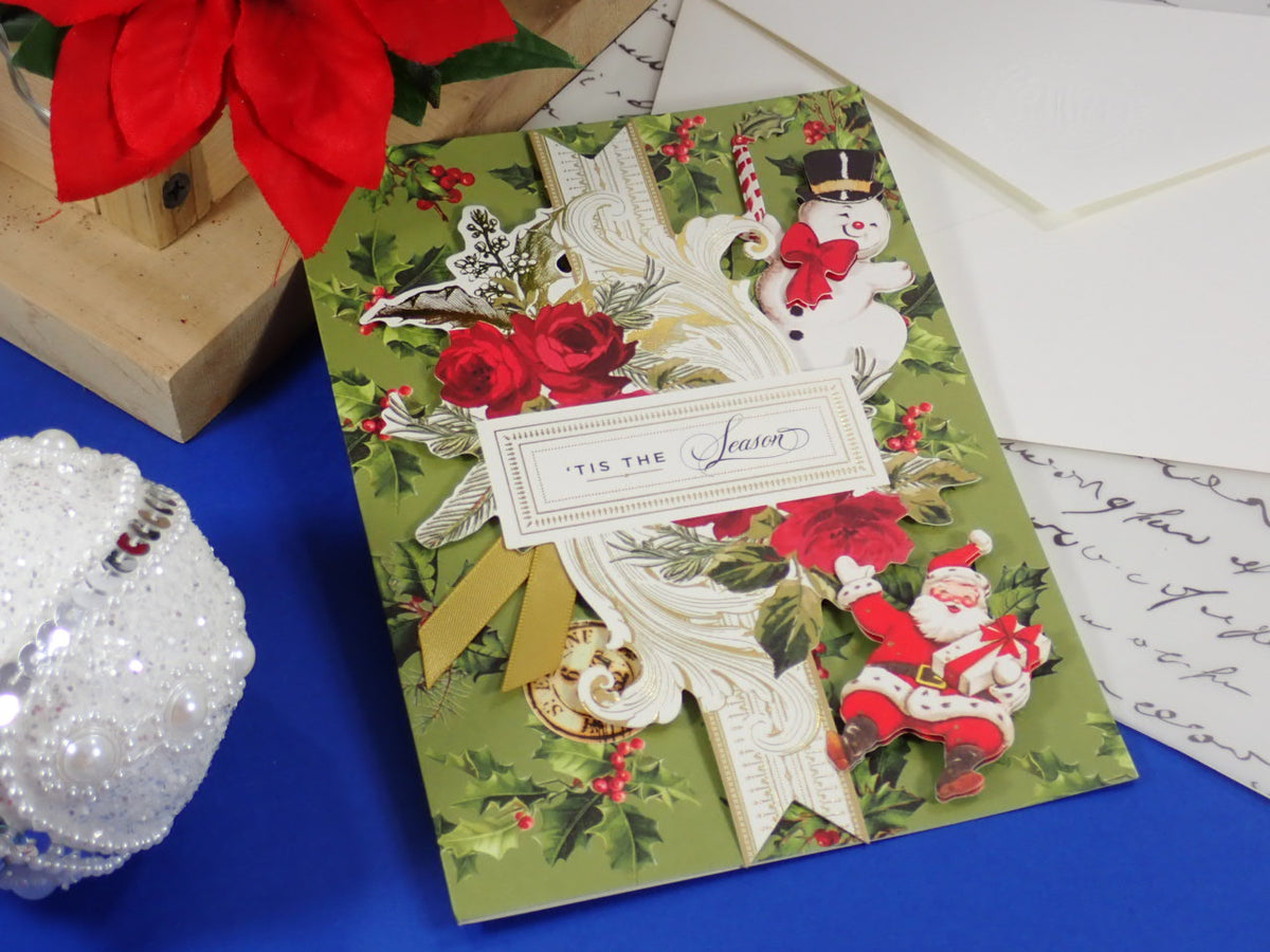 A handcrafted Christmas greeting card with a vintage holiday design, featuring a green background, red roses, snowman and Santa illustrations, and a white ribbon with a label that reads 'TIS THE Season'. 