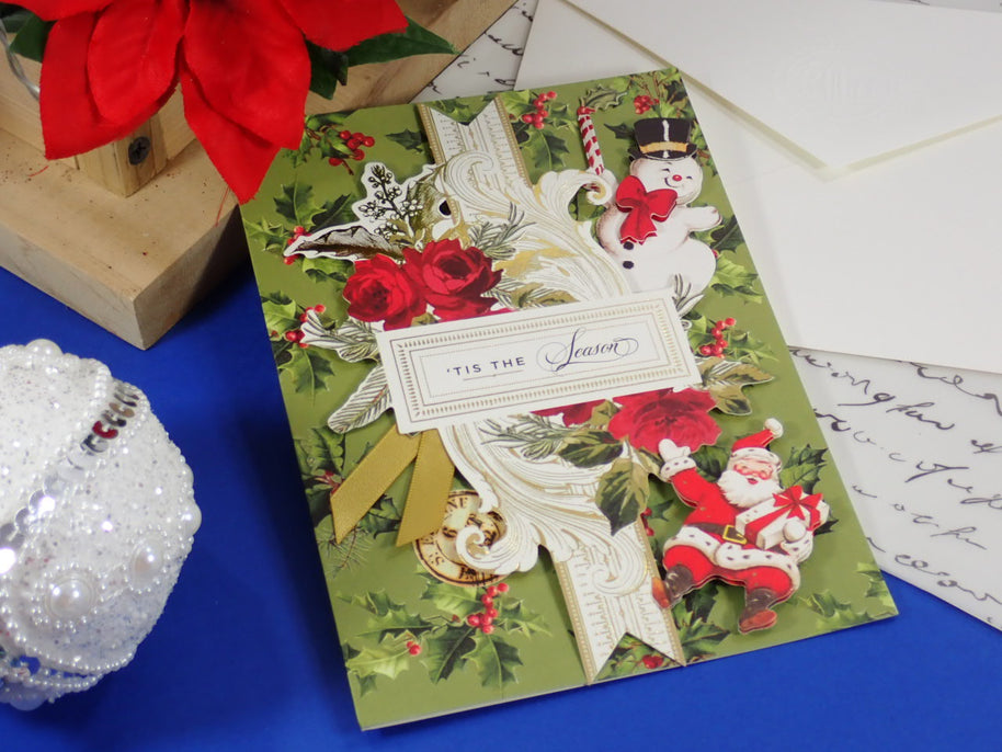 A handcrafted Christmas greeting card with a vintage holiday design, featuring a green background, red roses, snowman and Santa illustrations, and a white ribbon with a label that reads 'TIS THE Season'. 