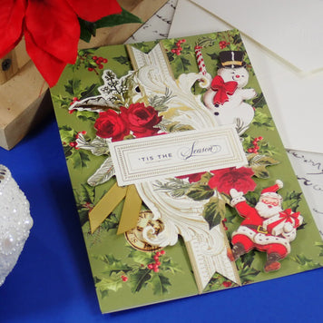 A handcrafted Christmas greeting card with a vintage holiday design, featuring a green background, red roses, snowman and Santa illustrations, and a white ribbon with a label that reads 'TIS THE Season'. 