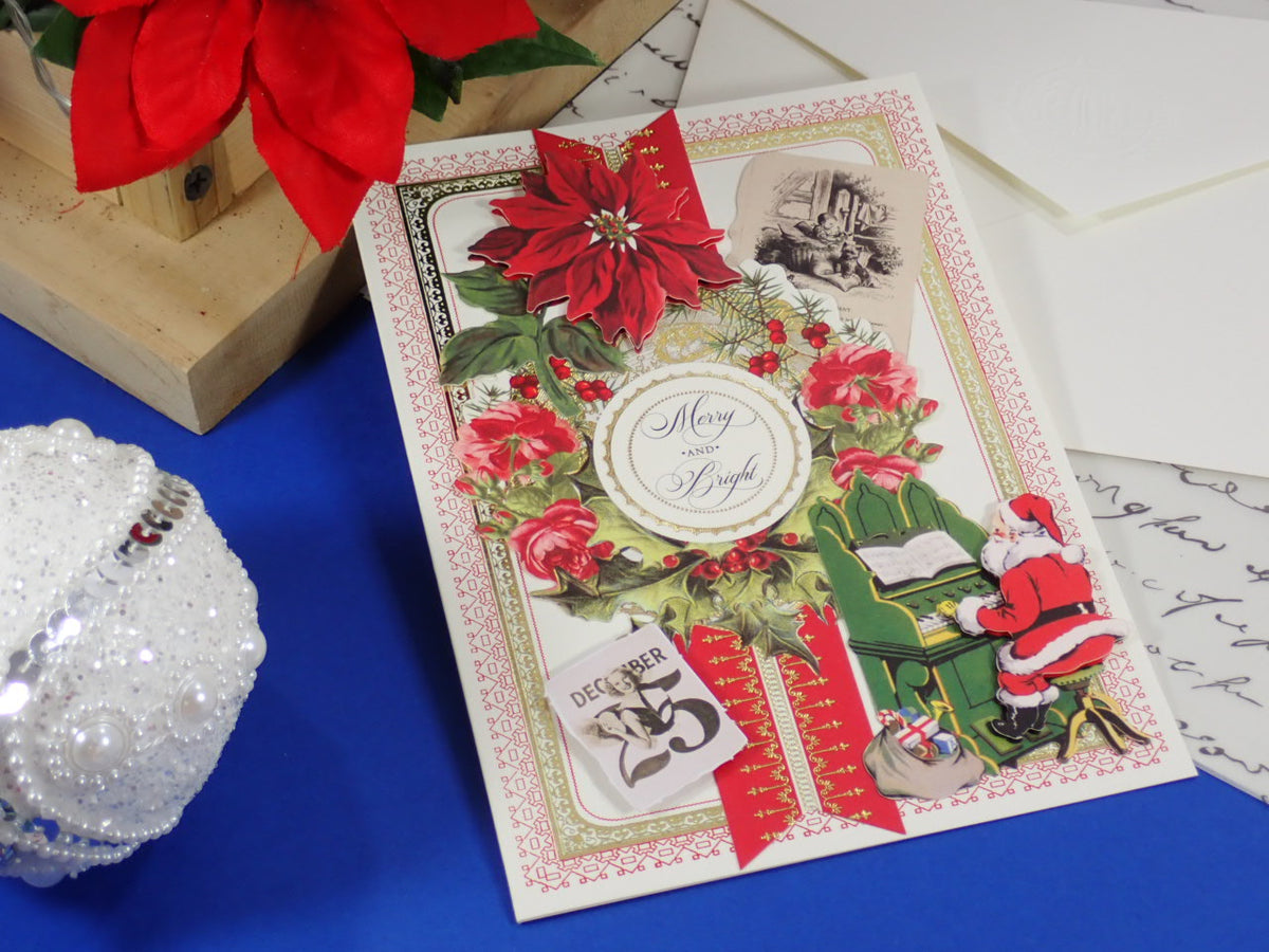 A handcrafted Christmas greeting card with red poinsettias, a Santa figure, and a central round label reading 'Merry and Bright,' styled on a blue surface with holiday props.