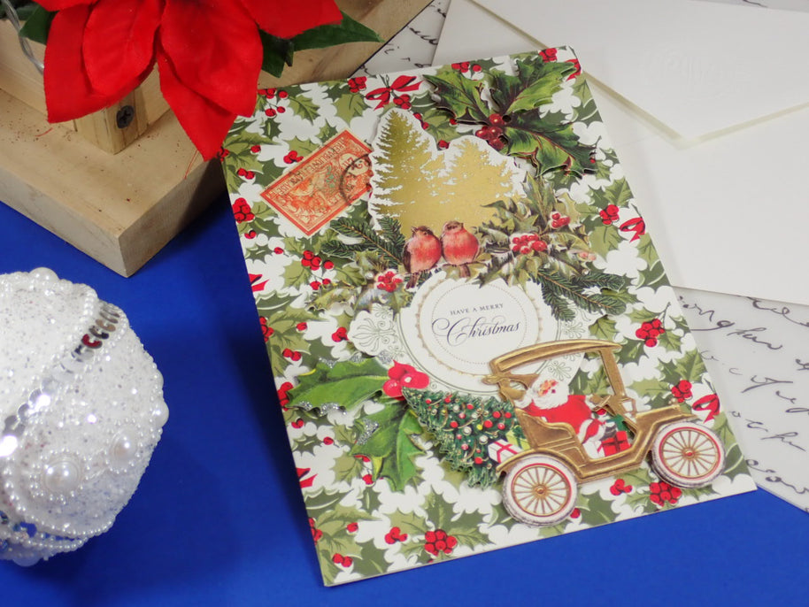 A handcrafted, Christmas greeting card featuring a vintage Santa Claus car, holly, birds, and a postage stamp design, styled on a blue surface with a white envelope and holiday decorations.