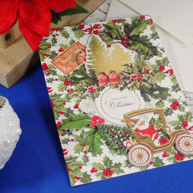 A handcrafted, Christmas greeting card featuring a vintage Santa Claus car, holly, birds, and a postage stamp design, styled on a blue surface with a white envelope and holiday decorations.