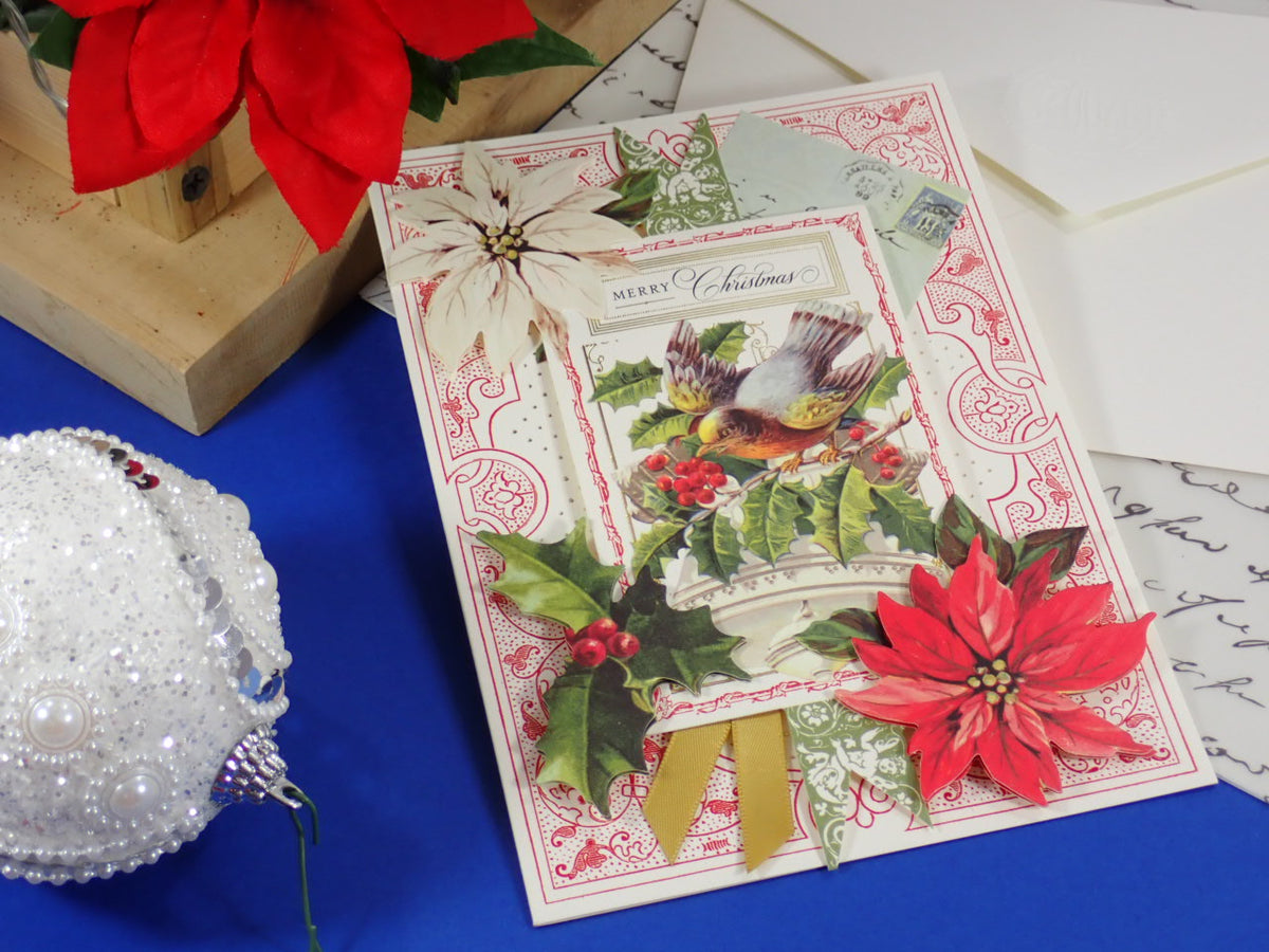 A handcrafted, vintage-style Christmas greeting card with a bird illustration, poinsettias, holly leaves, and a gold ribbon, styled on a blue surface with holiday decorations.