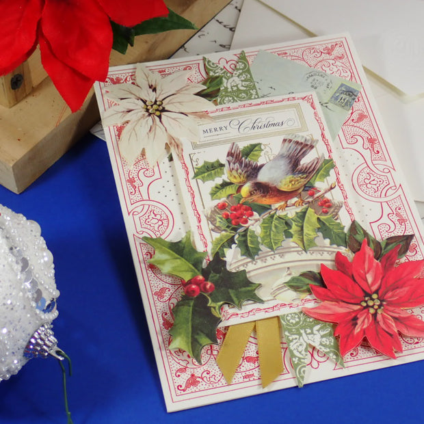 A handcrafted, vintage-style Christmas greeting card with a bird illustration, poinsettias, holly leaves, and a gold ribbon, styled on a blue surface with holiday decorations.