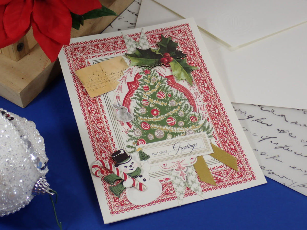 A handcrafted, vintage-style Christmas greeting card with a decorated tree, holly, snowman, and candy cane embellishments, displayed on a blue surface with white envelopes and holiday props around it.
