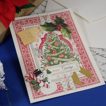 A handcrafted, vintage-style Christmas greeting card with a decorated tree, holly, snowman, and candy cane embellishments, displayed on a blue surface with white envelopes and holiday props around it.