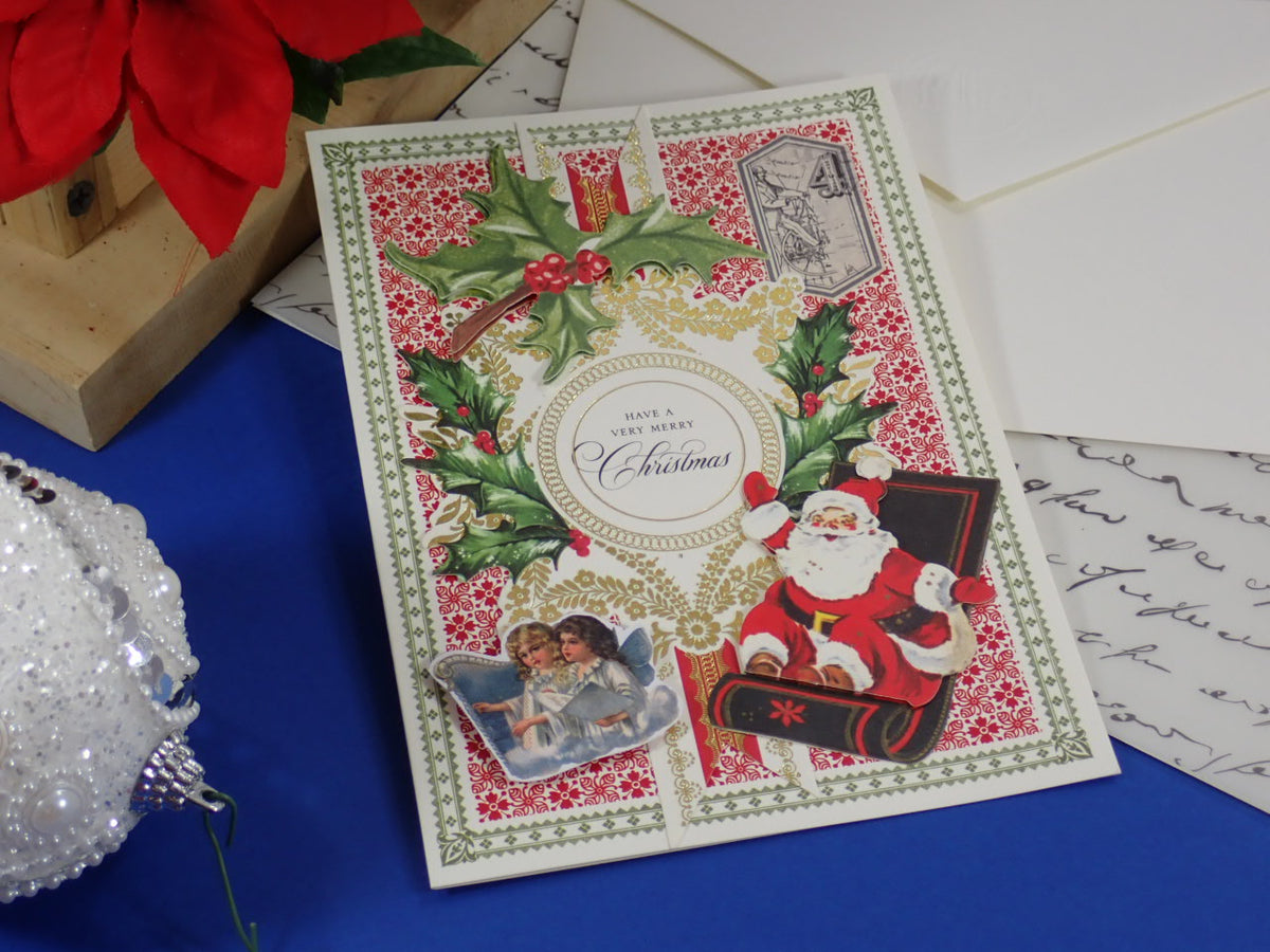A handcrafted Christmas greeting card with a vintage Santa Claus illustration, holly, red patterned background, and a central oval reading 'Merry Christmas.'