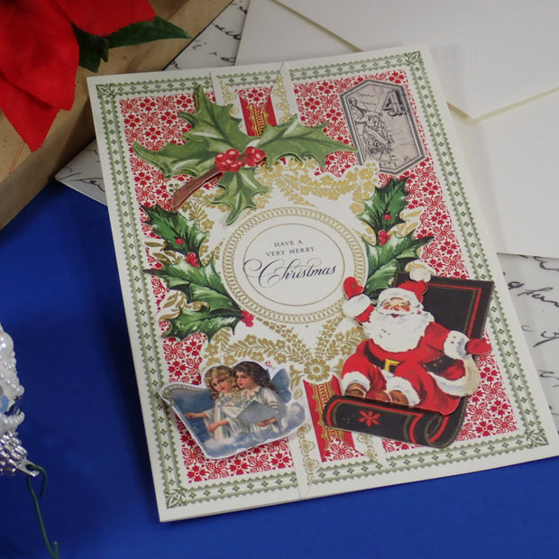 A handcrafted Christmas greeting card with a vintage Santa Claus illustration, holly, red patterned background, and a central oval reading 'Merry Christmas.'