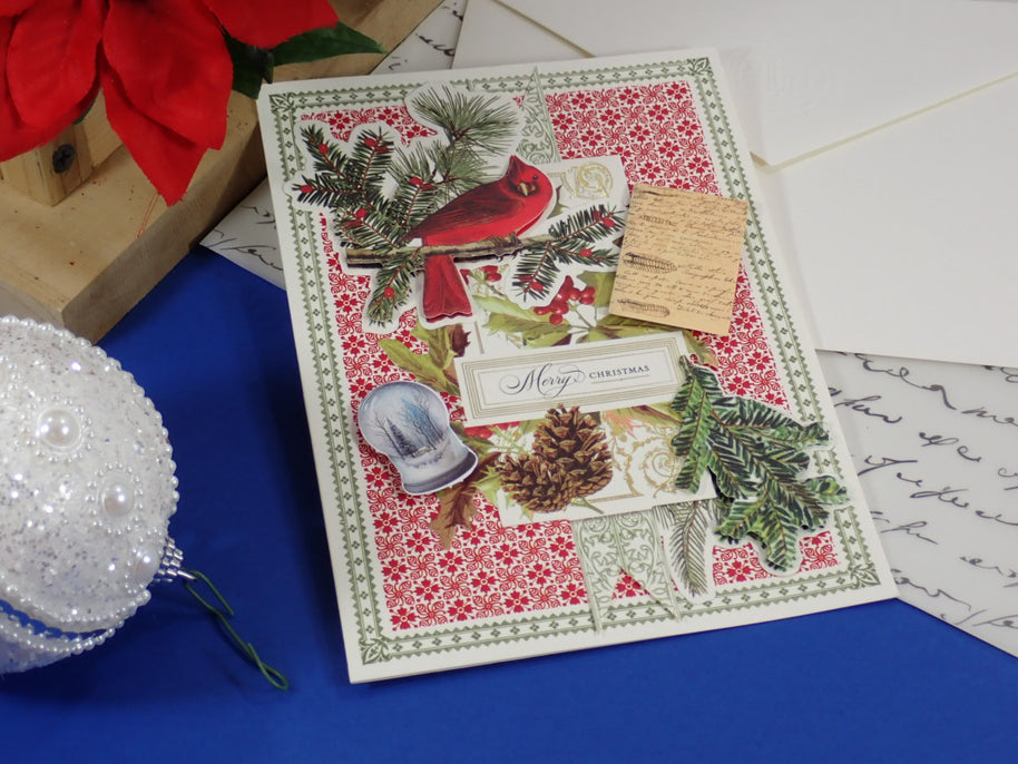 A handcrafted Christmas greeting card featuring a red cardinal, green pine branches, a red and white floral pattern, and a small wooden tag, styled on a blue surface with holiday decorations.