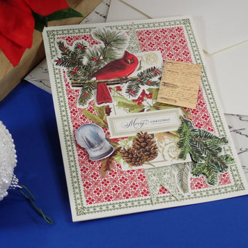 A handcrafted Christmas greeting card featuring a red cardinal, green pine branches, a red and white floral pattern, and a small wooden tag, styled on a blue surface with holiday decorations.