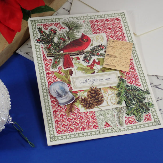 A handcrafted Christmas greeting card featuring a red cardinal, green pine branches, a red and white floral pattern, and a small wooden tag, styled on a blue surface with holiday decorations.