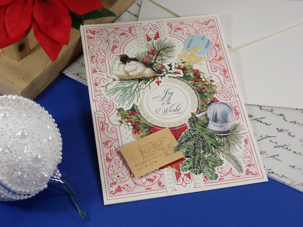 A handmade, vintage-style Christmas greeting card with ornate red and cream patterns, holly, pine branches, a bird illustration, and a small envelope, styled on a blue surface with holiday props.