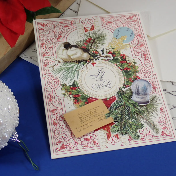 A handmade, vintage-style Christmas greeting card with ornate red and cream patterns, holly, pine branches, a bird illustration, and a small envelope, styled on a blue surface with holiday props.