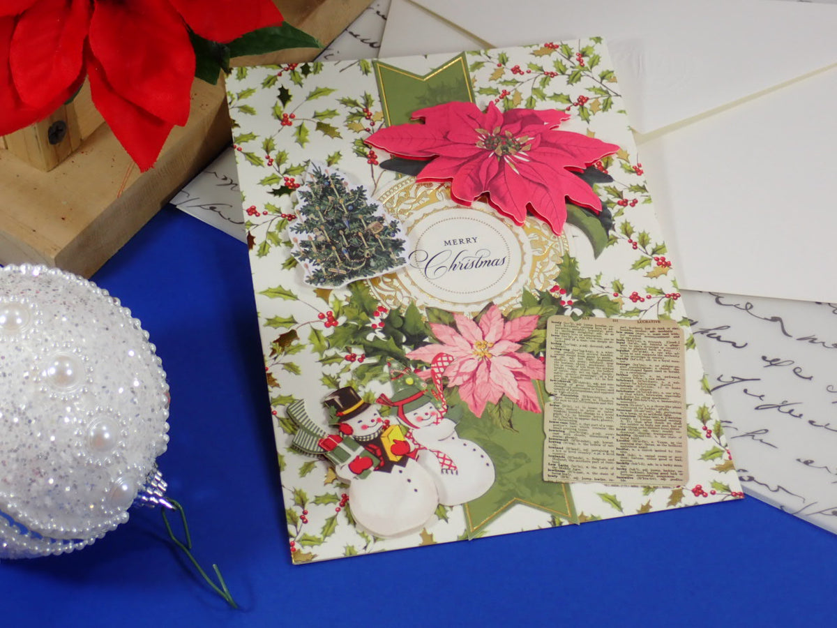 A handmade Christmas greeting card with a festive collage design, featuring pink poinsettias, green holly, snowmen, a small tree, and a vintage-style paper insert, displayed on a blue surface with holiday props.