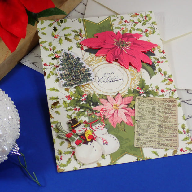 A handmade Christmas greeting card with a festive collage design, featuring pink poinsettias, green holly, snowmen, a small tree, and a vintage-style paper insert, displayed on a blue surface with holiday props.